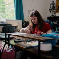 Nicola Allen works at her sewing table. It's got numerous tools used in sewing. She is focused.