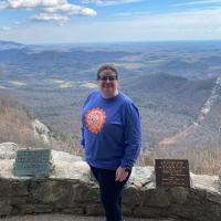 Nicola Allen, wearing a blue long-sleeved tee shirt with a green gear that says "Gear Up For Success," standing for a photo in front of a mountain overlook area.