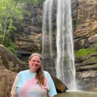 Nicola Allen is close to the foreground of a photo, wearing a tie-dye shirt. She's standing in front of a small pond being filled by a small waterfall.