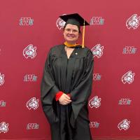 Nicola Allen dressed in graduation gown. She's smiling gleefully in front of a banner that indicates she is at Indiana Wesleyan University.