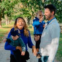 Marya Riggs, left, laughs while holding a toddler. Kyle Riggs smiles toward them while a toddler he's holding points their finger at his face.
