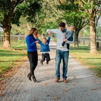 The Riggs family walks on a road on a farm. One of their two toddlers is being swung by the parents, who are are each holding a hand. The other toddler is in Kyle Riggs' left arm.