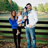 The Riggs family stands in front of a fence for a tall portrait shot. wife Marya is to the left of frame holding one daughter, Kyle Riggs is right in frame holding the other.