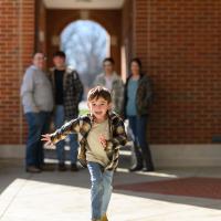 Granger Smith, very prominent in the foreground, runs toward the camera lens offscreen. His family is visible, though out of focus, in the background near a brick building.