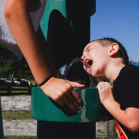 Granger Smith, right of frame, drinks from a water fountain as his mom, visible partially in the frame to the left, holds the water fountain activator button