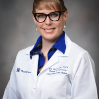 Dr. Erin Camac poses against a gray background wearing a white medical coat with her name written on it.