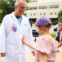 Scottie Day smiling, wearing a coat with a child's handprint painted on it, as a child with an IV in their face sticks a hand to his stomach