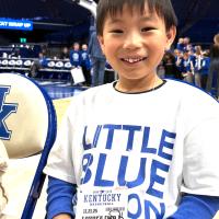 Child wearing a “Little Blue Nation” shirt holds a UK basketball game pass while standing courtside near the hoop during a game-day experience.