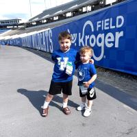 Two children wearing UK shirts and guest credentials stand together near Kroger Field signage during a football game-day visit.