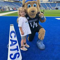 Child stands beside the UK Wildcats mascot on the Kroger Field turf during a game-day experience, with stadium seating visible behind them.