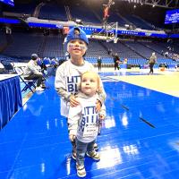 Two children wearing “Little Blue Nation” shirts stand courtside on the basketball floor before a UK game, with the hoop and arena seating in view.