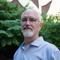Dan Gutenson poses for a portrait, wearing a polo, against foliage