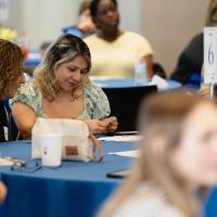 Maria Fuentes, in focus along with a friend, seated at a table observing something she's holding