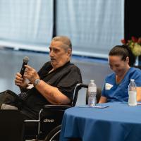 Jerry Gumm, holding a microphone, speaking from his table seated next to a friend who is laughing