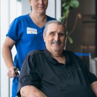 Jerry Gumm, seated in a wheelchair and smiling at camera, with an aide dressed in blue scrubs standing behind him