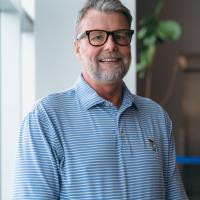 Michael Hudkins, smiling at camera, dressed in a blue striped polo and wearing thick glasses