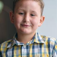 Child Oliver Blair, wearing a flannel-colored shirt, smiles at camera
