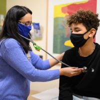 Dr. Preeti Ramanchandran, a middle-aged woman with black hair, glasses, a purple sweatshirt and blue mask, places a stethoscope on AJ’s chest as he sits facing her.