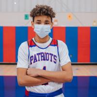 AJ stands in the middle of the basketball court in his white Patriots team uniform and a face mask. He has his arms crossed in front of his chest.