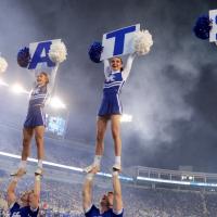 Allie and her fellow female UK cheerleaders smile as they hold blue and white pom poms and signs which read “C-A-T-S.” They are being held up high by the male cheerleaders.