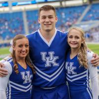 Allie smiles and poses with two other UK cheerleaders on a football field.