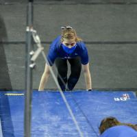 Allison squats to position a mat in place at a gymnastics practice. She is wearing a blue facemask, blue tee shirt, and black leggings. Her hair is pulled back and has a French braid across the front.