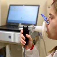 Allison sits in an exam room and conducts a lung test, blowing into a hand-held piece of equipment while her nose is blocked with a blue clip.