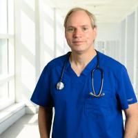 Dr Michael Anstead, a middle-aged white man with blonde hair, stands smiling in a white hallway. He is wearing blue scrubs and has a stethoscope around his neck.