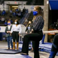 In a black and white blouse, black slacks, and a blue facemask, Allison stands at the edge of a competition space and watches her gymnastics team compete.