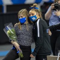Allison poses for a photo with a fellow gymnastics teammate. They have their arms around each other. Allison is holding a bouquet of flowers. Both are wearing blue facemasks.