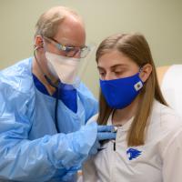Dr. Anstead places a stethoscope on Allison’s chest during an exam. He is wearing a blue gown over his blue scrubs, a face mask, face shield, and gloves. Allison is wearing a white UK athletics top and a blue facemask.