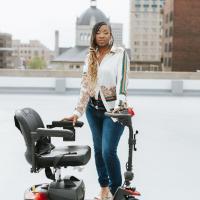 A photo of Brandi standing as she poses with her electric scooter. She is on the roof of a building, and many more can be seen behind her.