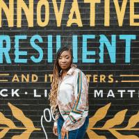A photo of Brandi standing and posing confidently in front of a mural that reads “Innovative Resilient And It Matters. Black Lives Matter.”