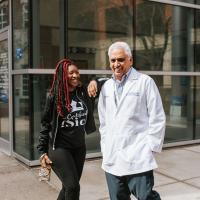 A candid photo of Brandi and Dr. Avasarala laughing together as they stand outside. Dr. Avasarala is an older South Asian man with short gray hair. He is wearing a white lab coat over a blue and white button-up shirt, and blue jeans.