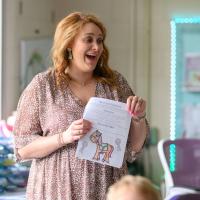 Callie, wearing a brown and white floral dress, stands in the middle of her elementary school classroom. She is laughing and looking off-camera, and is holding a worksheet with a horse on it.