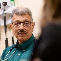 Dr. Jamshed Kanga, an older Indian man with short gray hair, glasses, and a mustache, talks with a patient in the extreme foreground of the photo. He is wearing a green collared shirt and has a stethoscope around his neck.