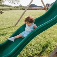 Charlie, dressed in a gingham overall set with horses on it, slides down a green slide attached to a wooden playground in the family’s backyard.