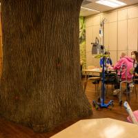 A candid photo of two UK HealthCare employees sitting with a patient in one of the Child Life rooms, which has a giant fake tree trunk in the middle of it. The patient is a white teenage girl with a bald head. She is wearing a pink hoodie.