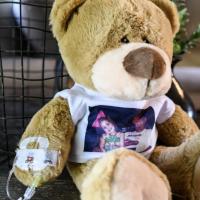 A photo of Henley Thurman’s stuffed bear. It has an IV in its arm, and is wearing a white t-shirt that has a picture of Henley on it.