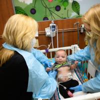 An over-the-shoulder shot of Jennifer (left) and Ashlee Olson (right) lifting up a young patient out of their bed. The patient has several monitors hooked up to them, as well as an oxygen tube attached to their nose. Ashlee is a young white woman with long blonde hair that has been curled. She is wearing a blue plastic cover over her clothes, a blue face mask, and round glasses.