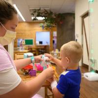 An over-the-shoulder shot of a mother playing kitchen with her child, a Child Life patient. The woman is a white woman with brown hair that is tied up in a bun. She is wearing a short-sleeve pink t-shirt over a long-sleeve white shirt, and a yellow face mask.