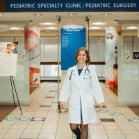Dr. Laura Stadler, a white woman with chin-length blonde hair, stands outside of the Pediatric Speciality Clinic inside UK HealthCare. She is wearing a doctor’s coat with a stethoscope around her neck.