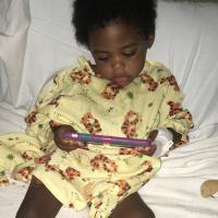 Jackson sits on his hospital bed, surrounded by his toys as he watches something on a phone. He is wearing a hospital gown with tigers on it.