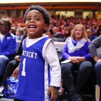 Jackson stands on the sidelines at a basketball game, smiling widely. There are several seated people behind him wearing blue and white UK colors. Jackson is wearing a Kentucky basketball jersey over a long-sleeve white shirt.