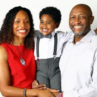 Jackson smiles widely as he is carried in between his mother, Coach Kyra Elzy—an African American woman with brown hair in a red dress—and his father, Dexter Lander, an African American man in a white button-down shirt