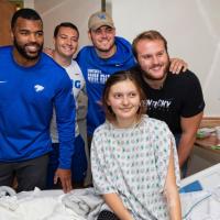 Courtney Love, wearing a blue shirt, and three current football players wearing Kentucky gear, pose with a Kentucky Children’s Hospital patient. The patient, a young girl, is sitting in her bed and wearing a hospital gown.