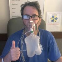 Dr. Johnson sits in a hospital room, with an oxygen mask over his nose and mouth. He is wearing a blue shirt, and giving a thumbs-up sign with his right hand.
