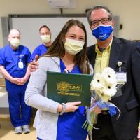 Holding a bouquet of flowers, Alexis Zody, an adult white woman with brown hair in blue scrubs, stands beside Dr. Johnson for a photograph. They are surrounded by colleagues, and everyone pictured is wearing a facemask.