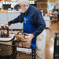 Donald is putting groceries in a plastic bag at a Kroger supermarket. He is wearing a facemask, a khaki baseball cap and a blue Kroger apron and nametag.
