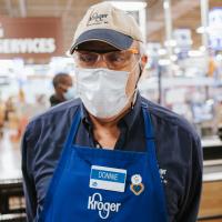 Donald standing in the Kroger supermarket where he works. He is wearing a brown and blue cap, a blue Kroger apron, his name badge, a facemask and transparent safety glasses. He is holding his hands behind him and appears to be smiling under his mask.